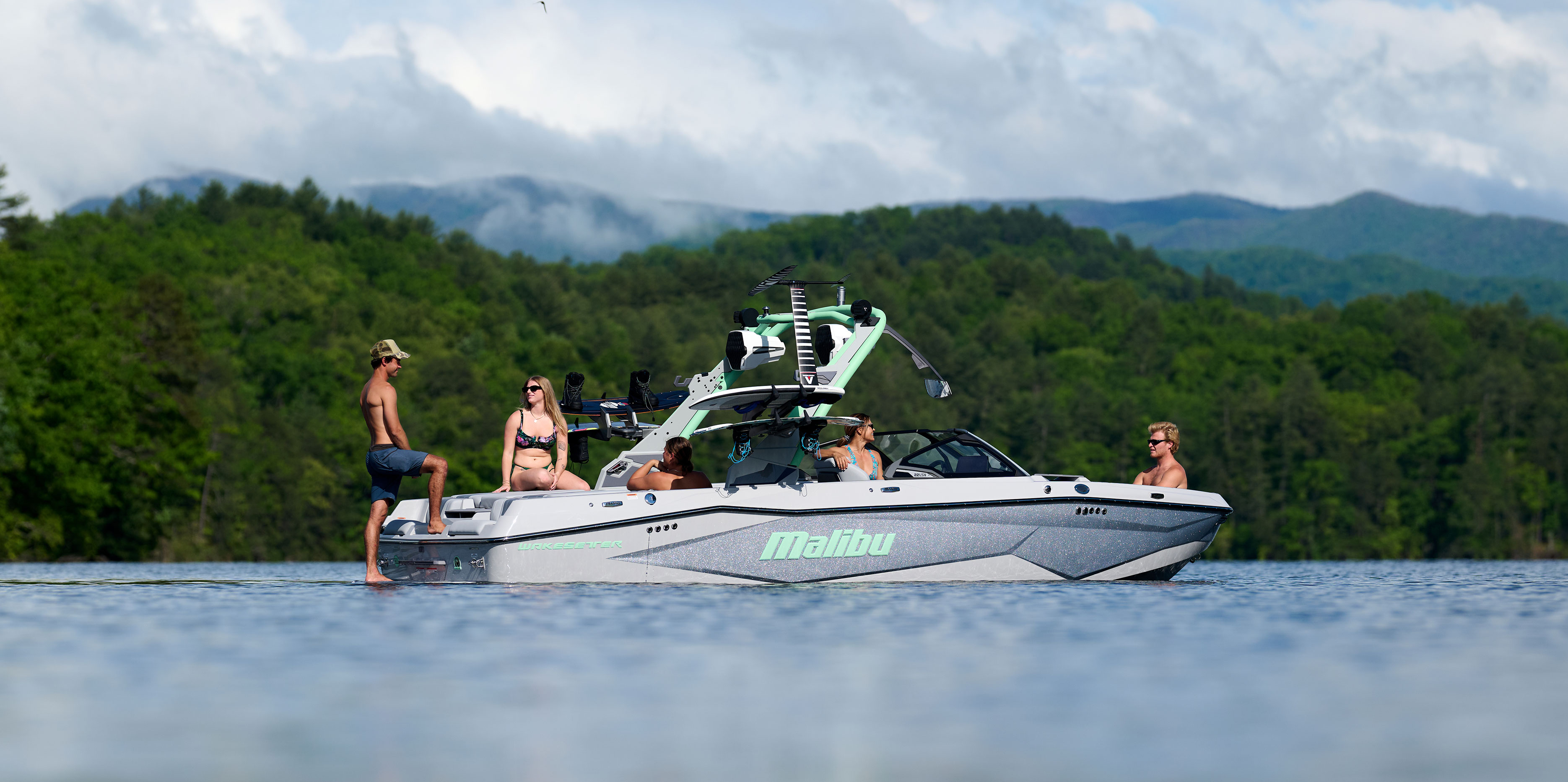 Group of people relaxing on the Malibu Wakesetter 22 LSV with the Ascend Tower, floating on a lake surrounded by forested mountains.
