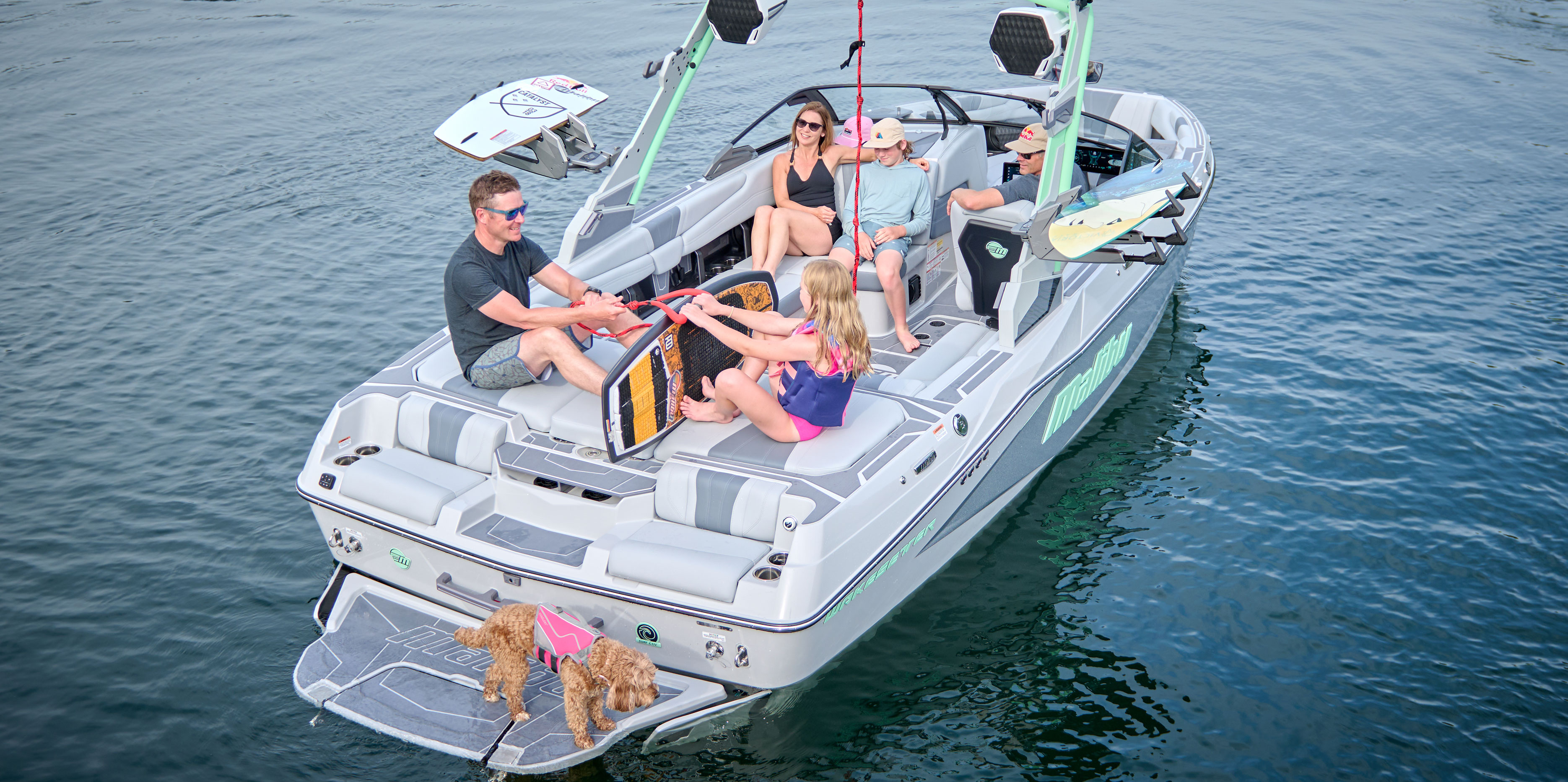 Wide-angle view of the Malibu Wakesetter 22 LSV with a surfer riding a clean wave and passengers enjoying the lake setting.