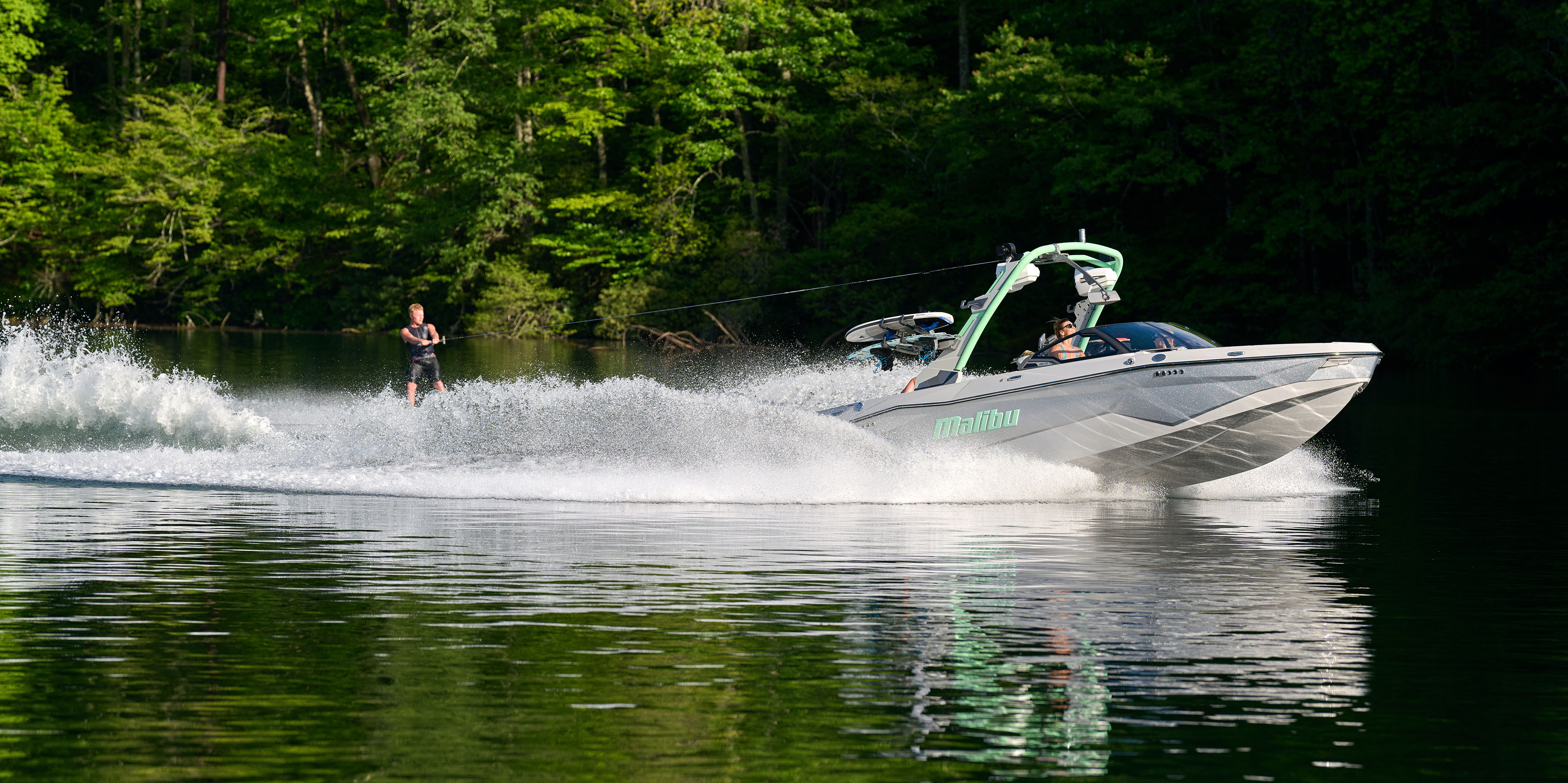Surfer rides a tall, clean wave behind the Malibu Wakesetter 22 LSV with vibrant water spray and scenic background.